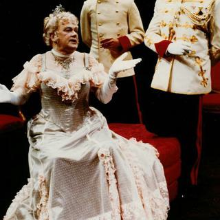 Three actors on stage, one is wearing a period ladies costume and seated with arms outstretched, the other two actors stand behind her.
