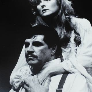A man is sat wearing a white shirt and braces, holding the hand of a woman standing behind him with her arms around his shoulders