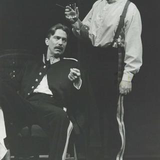 A man sits in military formal wear, whilst another man in a white shirt, braces and trousers stands behind him talking with a drink in his hand