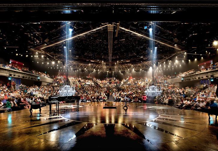 The Festival Theatre auditorium is seen from the stage. The audience is full. The black and gold stage stretches out in front of the camera. On the left of the frame is a grand piano. Two white crystal chandeliers hang down from the ceiling. There are lots of golden lights shining towards the stage and the camera from the ceiling