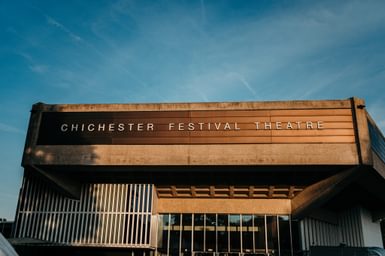 We're looking up at the listed Festival Theatre building, a brutalist design, against a clear blue summer's evening sky. The theatre appears to be lit by late afternoon sun and appears almost gold in colour.