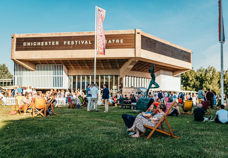 A full lawn of people dressed in summer clothes are enjoying drinks, chatting and milling around outside the Festival Theatre. We can see the building clearly in the background against a bright blue summer sky and there are multicoloured flags flying in front of it which say 'Festival 2023' on them.