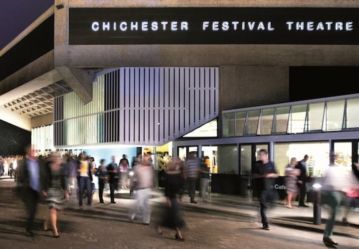 The outside of the Festival Theatre at night time. The building is lit and there is a blur of people moving around outside.