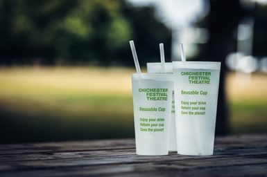 Two of our frosted reusable cups with green writing on the side and green striped paper straws sticking out of them.