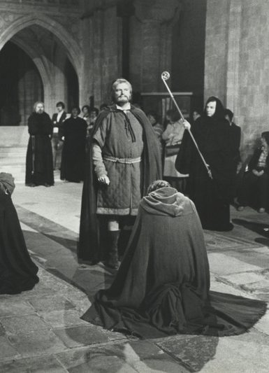 Black and white scene of actors in long robes performing a dramatic moment inside a large stone cathedral.
