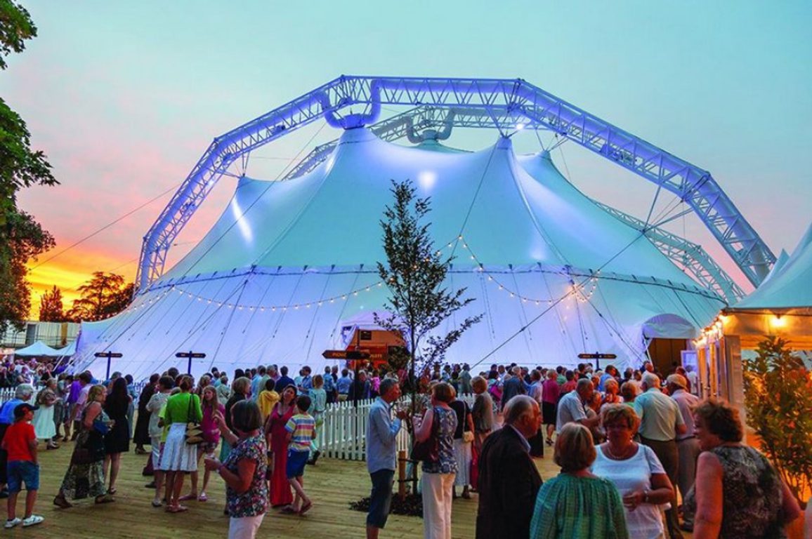 A large outdoor tent lit in soft blue at sunset with a crowd gathered around the entrance.