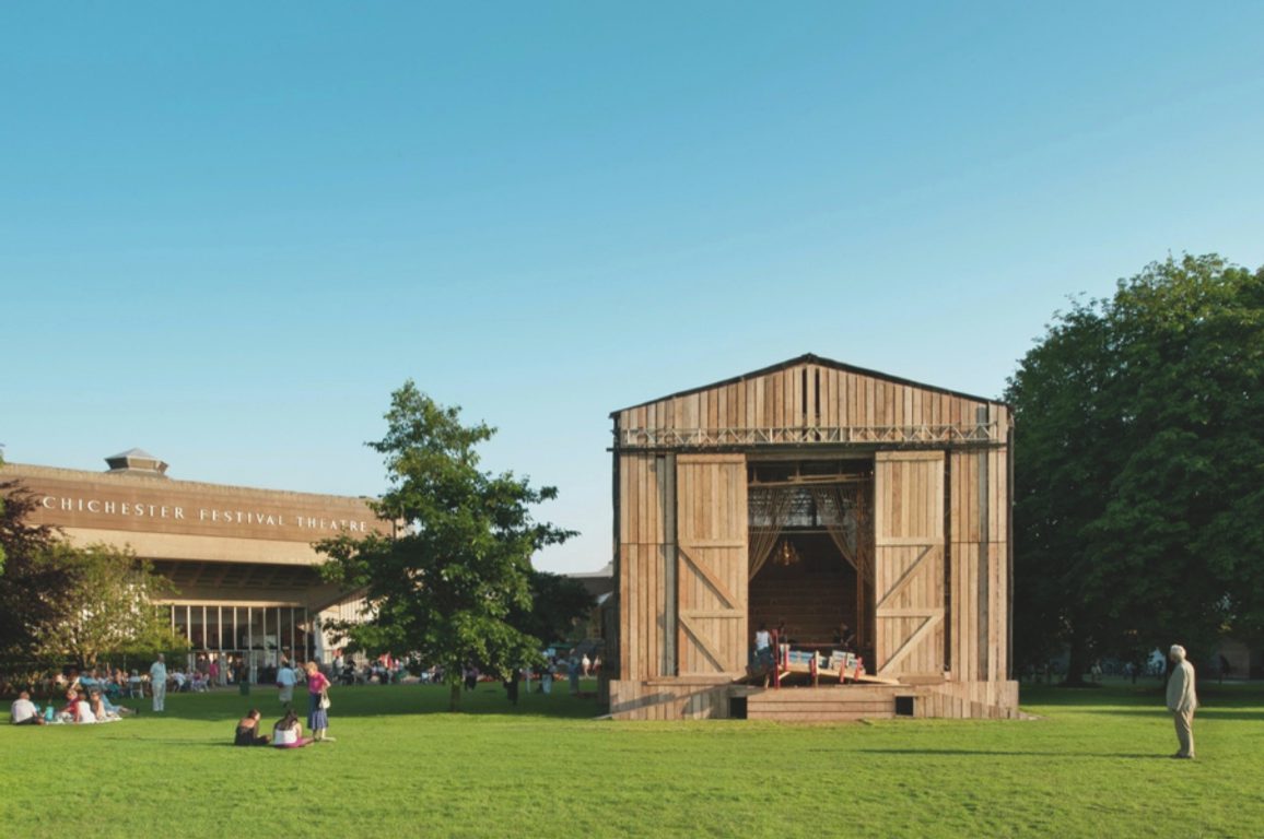 A wooden open‑air stage standing on a wide green lawn with people relaxing nearby under a clear sky