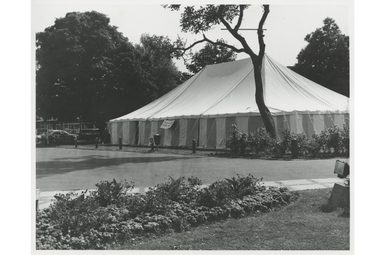 A large striped tent set up in a park with trees, bushes, and a flower bed in the foreground, captured in a black and white photo.