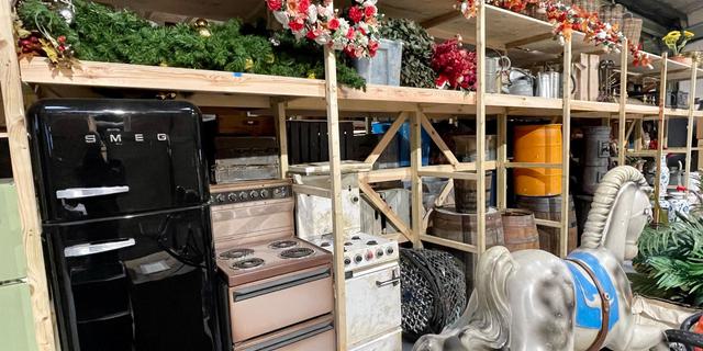 A variety of items including a black SMEG fridge, two vintage stoves, a piebald rocking horse, wooden barrels and baskets of artificial flowers are piled on and beside high wooden shelves