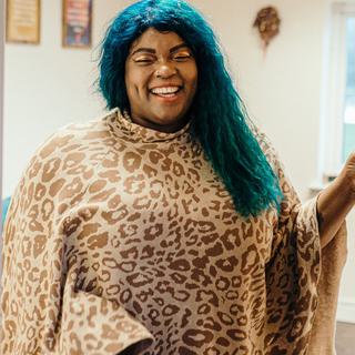 Shari, the CFT creative therapist, is stood in a doorway. She is smiling at the camera with her hand on the door. Her hair is turqouise, and she is wearing a leopard print poncho.