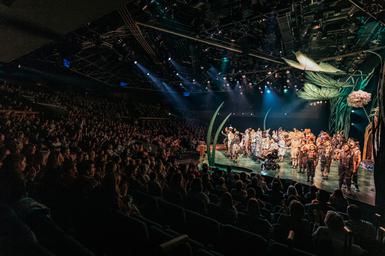 A photo from the side of the stage looking out over a full audience, applauding. The youth theatre company of Wind in the Willows are taking their bows on a colourfully lit stage.