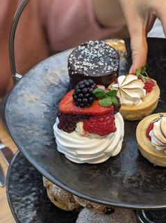 Afternoon tea on a tiered platter - mini cakes on a grey plate and a woman's hand is picking up a small cream cake.