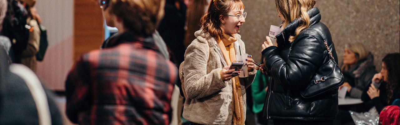 Two audience members stand in the Festival Theatre foyer. They are holing drinks and their phones. The person on the left is wearing a beige coat and has red hair. They excitedly smile at the person in front of them, whose face is obscured. Another group stands closer to the camera with their faces obscured.