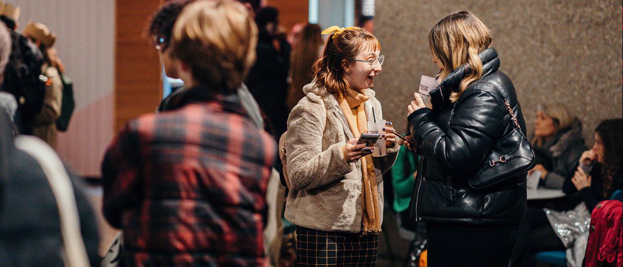 Two audience members stand in the Festival Theatre foyer. They are holing drinks and their phones. The person on the left is wearing a beige coat and has red hair. They excitedly smile at the person in front of them, whose face is obscured. Another group stands closer to the camera with their faces obscured.