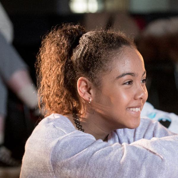 Three young people sit, smiling in a rehearsal rooml. They are wearing hoodies and jumpers.