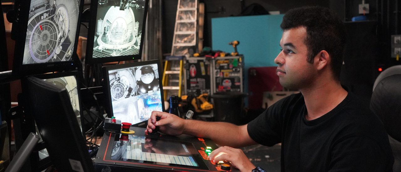 A young apprentice sits at a technical desk. He looks up at several screens with a focused expression.