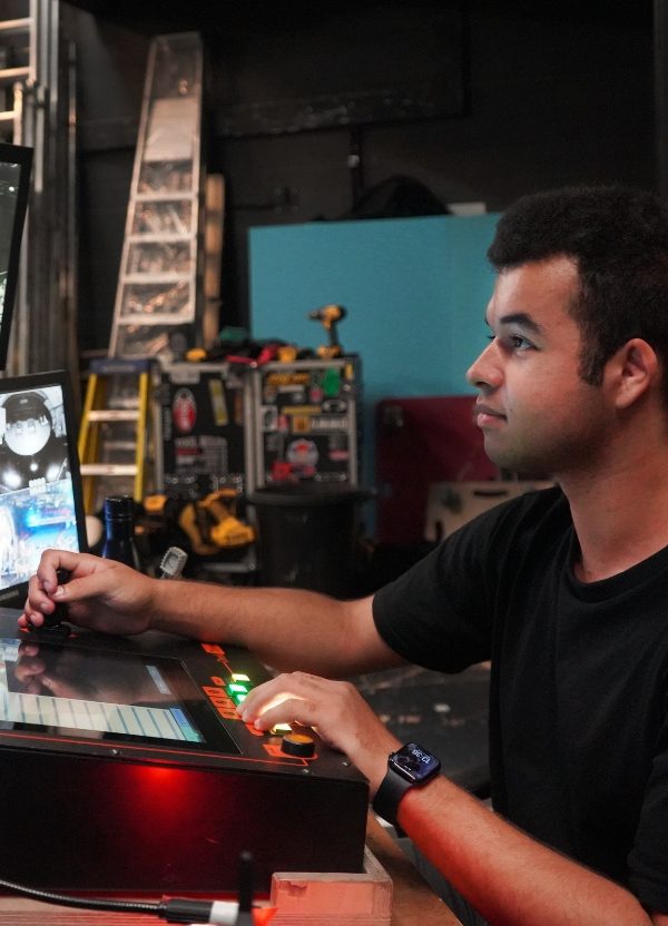 A young apprentice sits at a technical desk. He looks up at several screens with a focused expression.