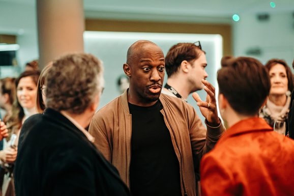 A man speaks enthusiastically to a group who stand around. In the background, others mingle.