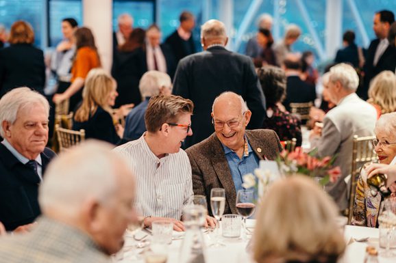 A group sit together at a formal dinner. In the centre, two guests laugh together.
