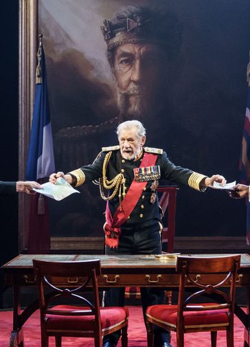King Lear stands centre, wearing military blues decorated with medals, behind a wooden table and chairs, holding out papers to two men in formal dress with their backs to the photographer. There is a large painting of the king looming over them on the wall, rich red carpets, and others in formal dress looking on from the sides.