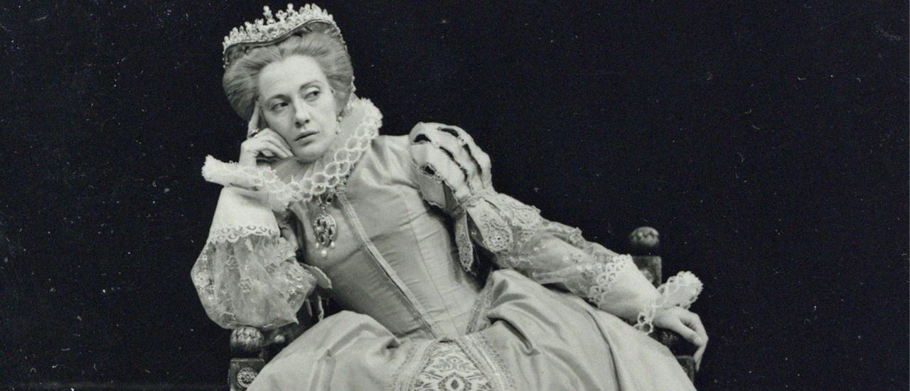 An actress dressed in a tudor costume sits and leans to one side, giving someone an evil side eye
