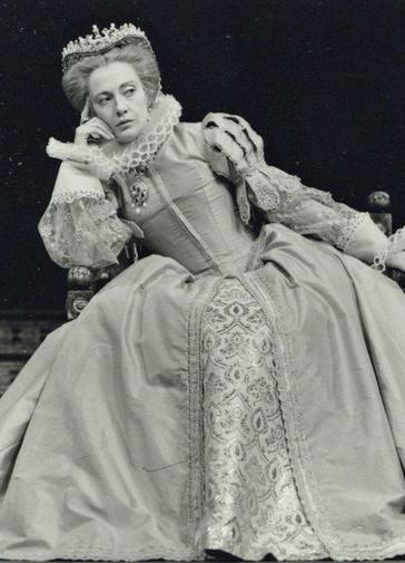 An actress dressed in a tudor costume sits and leans to one side, giving someone an evil side eye