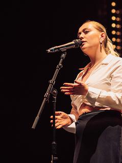 A young woman with blonde tied-back hair in a white blouse stands on stage. She is singing into a microphone, with her hands in front of her, her palms open in an expressive gesture. Behidn her are twinkly golden lights.