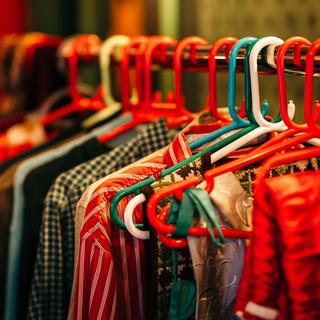 A clothing rack with colourful red, white and green hangars, it holds a row of Santa's clothes. In focus there is a pair of folded red trousers, a stripy red shirt, and a checked green and white shirt.