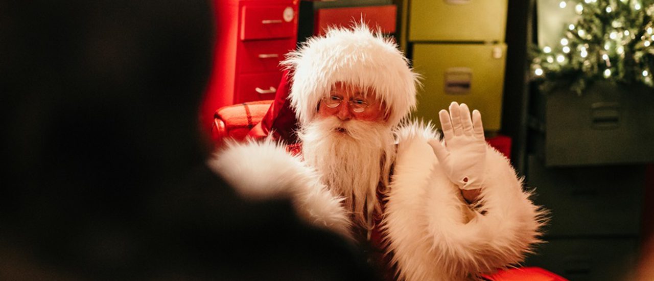 Santa waves from his red arm chair. He wears small spectacles, white gloves, a large red coat with fluffy white trims and has a long fuzzy white beard. Behind him is an assortmenyt of colourful drawer units, one drawer is open and a christmas tree rises out of it with warm lights on its branches.
