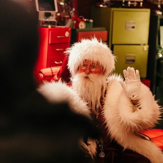 Santa waves from his red arm chair. He wears small spectacles, white gloves, a large red coat with fluffy white trims and has a long fuzzy white beard. Behind him is an assortmenyt of colourful drawer units, one drawer is open and a christmas tree rises out of it with warm lights on its branches.