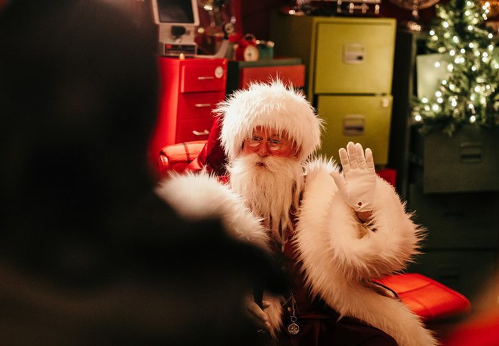 Santa waves from his red arm chair. He wears small spectacles, white gloves, a large red coat with fluffy white trims and has a long fuzzy white beard. Behind him is an assortmenyt of colourful drawer units, one drawer is open and a christmas tree rises out of it with warm lights on its branches.