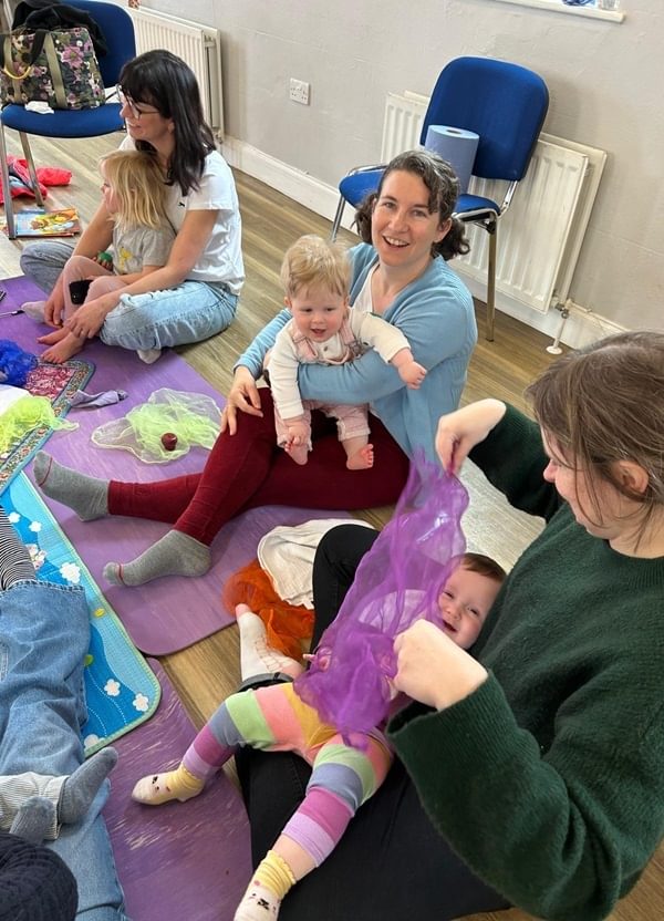 Mums and baby sit playing on the floor with silk scarves and toys.