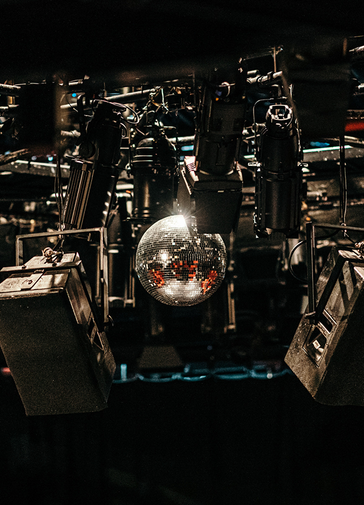 A glitterball shines and sparkles in the centre of the frame, with stage lights aimed at it. Below and beside it, stage speakers are hung.