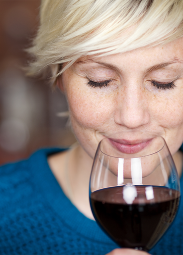A headshot of a lady in a blue jumper smelling the aroma of a glass of red wine.