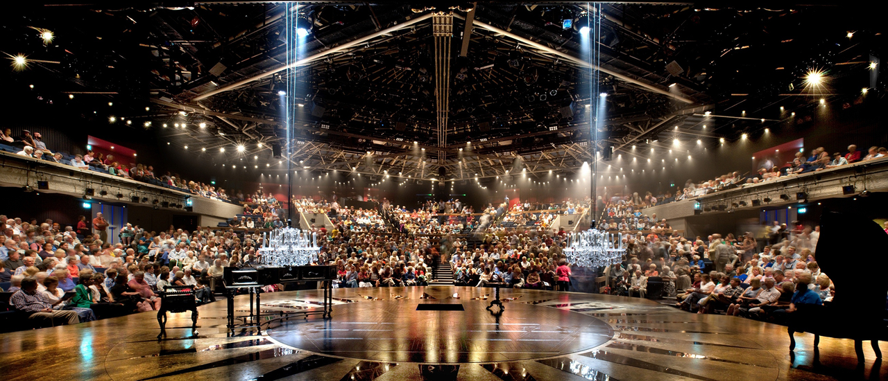 The Festival Theatre auditorium is seen from the stage. The audience is full. The black and gold stage stretches out in front of the camera. On the left of the frame is a grand piano. Two white crystal chandeliers hang down from the ceiling. There are lots of golden lights shining towards the stage and the camera from the ceiling
