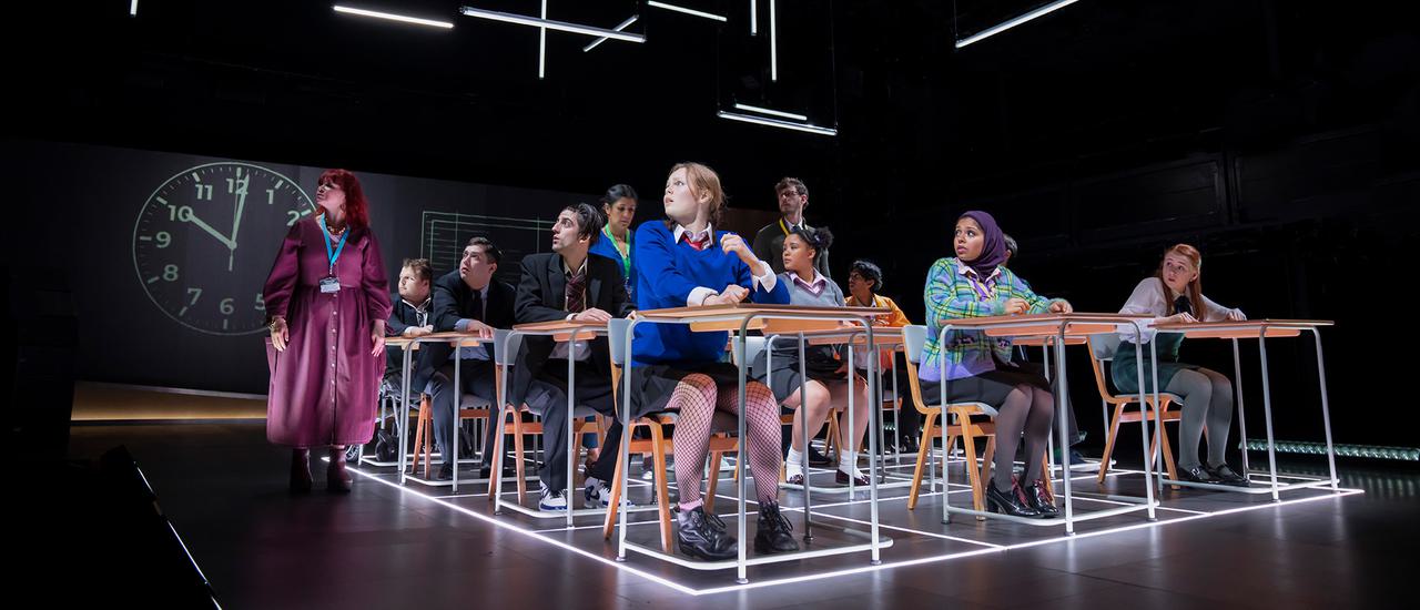 A school classroom with three rows of desks stretching back. Pupils are sitting at the desks, all looking to their right past a teacher on their right, who also looks in the same direction. There is a large clock projected on the back wall and strip lights arranged at various angles above their heads.