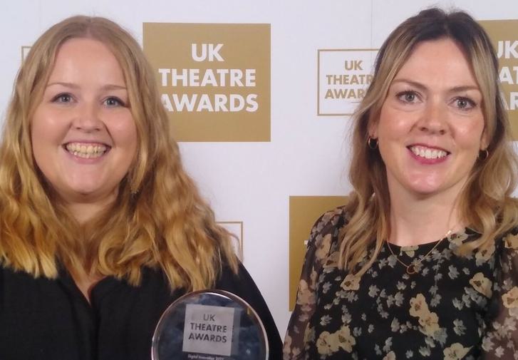 A woman in a black top and a woman in a flowery top stand in front of a banner displaying the UK Theatre Awards logo. They are smiling and holding an award.