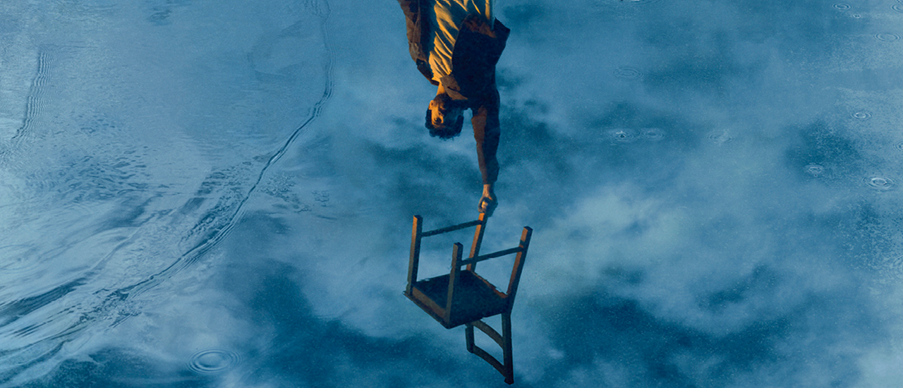 A reflection of sky in water. In the centre, a man with his hand stretched out is upside down holding a chair.