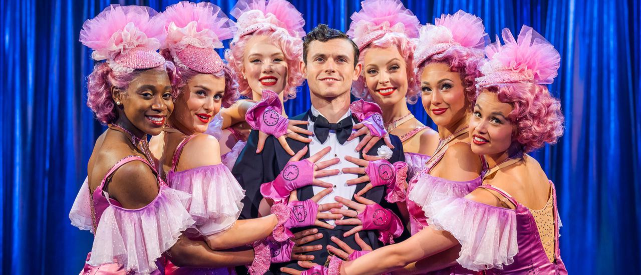 Bobby (Charlie Stemp) is dressed in a black suit with a white shirt on, he stands smiling surrounded by Follies dressed in pink frilly dresses and fascinators. They all have their hands on him.