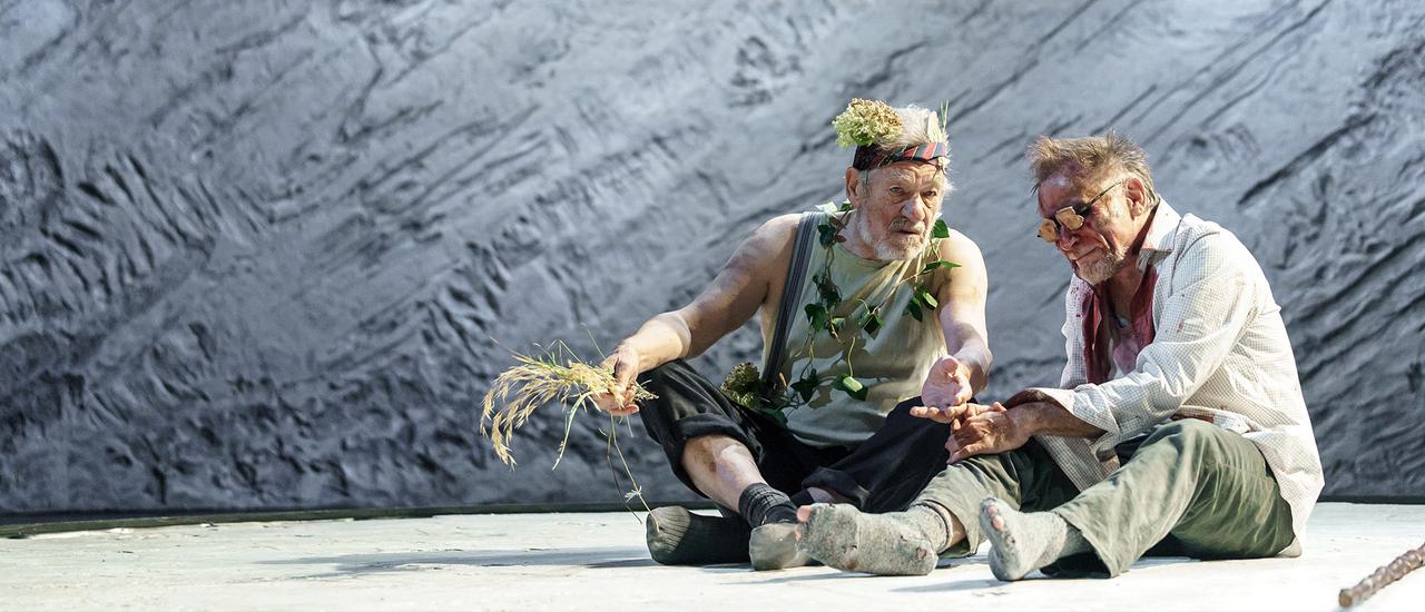 Two older men are sitting on a white floor, there is a grey background. Ian McKellen, the man on the left playing King Lear is holding some dried flowers and wearing flowers around his neck and head. He looks bedraggled. The man on the right, Danny Webb playing the Earl of Gloucester also looks bedraggled and has dried blood around his eyes which are covered with obscured glasses.