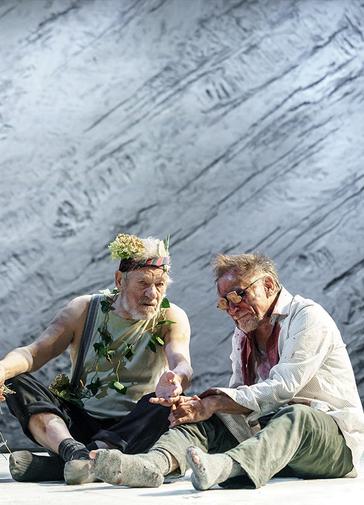 Two older men are sitting on a white floor, there is a grey background. Ian McKellen, the man on the left playing King Lear is holding some dried flowers and wearing flowers around his neck and head. He looks bedraggled. The man on the right, Danny Webb playing the Earl of Gloucester also looks bedraggled and has dried blood around his eyes which are covered with obscured glasses.