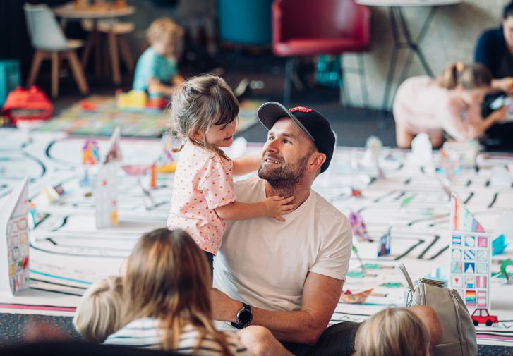 A father and daughter happily playing together in the theatre foyer surrounded by other families. Behind them you can see the outline of a large map of Chichester on the floor, being decorated by the families.