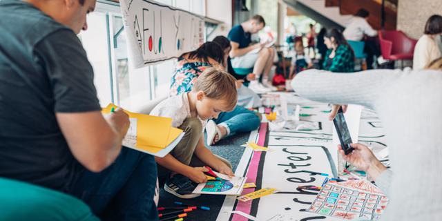Parents and children sit against the windows in the CFT foyer with apper and pens. The walls and floor are covered in crafts.