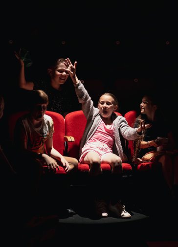 A girl is sat in a red theatre seat, with her hands held out dramatically. She is in a spotlight. Around her in the shadows are other smiling children.