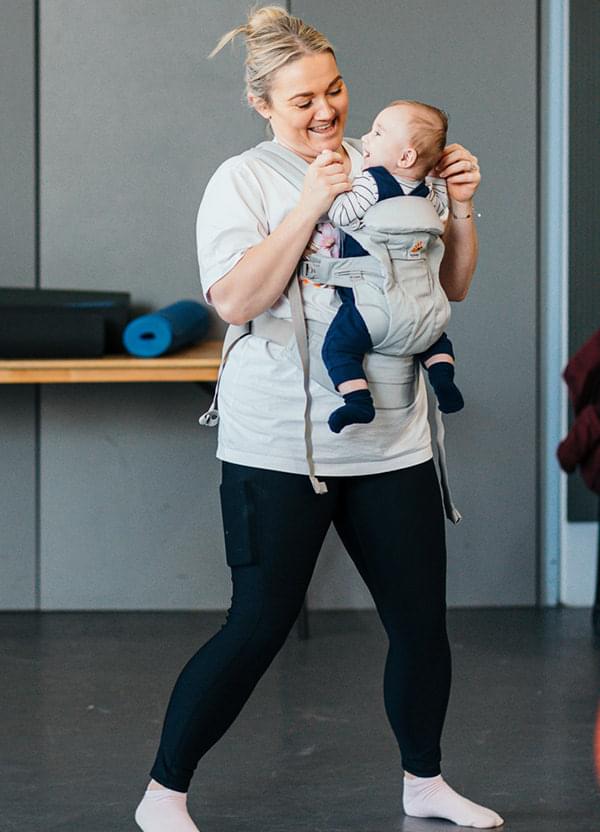 A woman and a little baby in a sling dance while looking and smiling at each other.