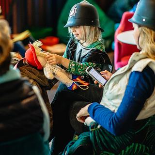 A girl in a plastic police hat plays with a soft doll. A woman, also in a plastic police hat sits next to her.