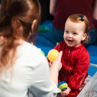 A smiling baby wearintg a red boiler suit sat in a box full of toys.