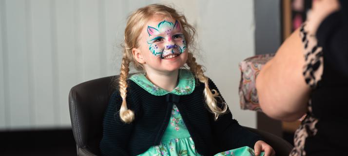 A young girl wearing a green dress and black cardigan is getting her face painted as a blue cat.