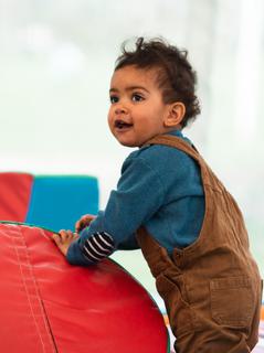A toddler wearing brown dungarees leaning against soft play equipment.