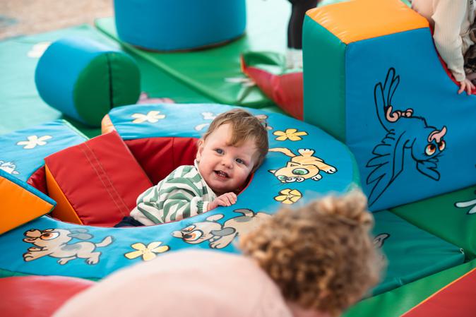A smiling baby in wearing a green and white stripy top is peeking over soft play equipment.